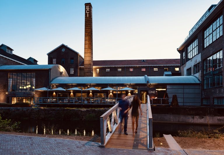 Restaurant in industrieel gebouw met schoorsteen, terras en brug.
