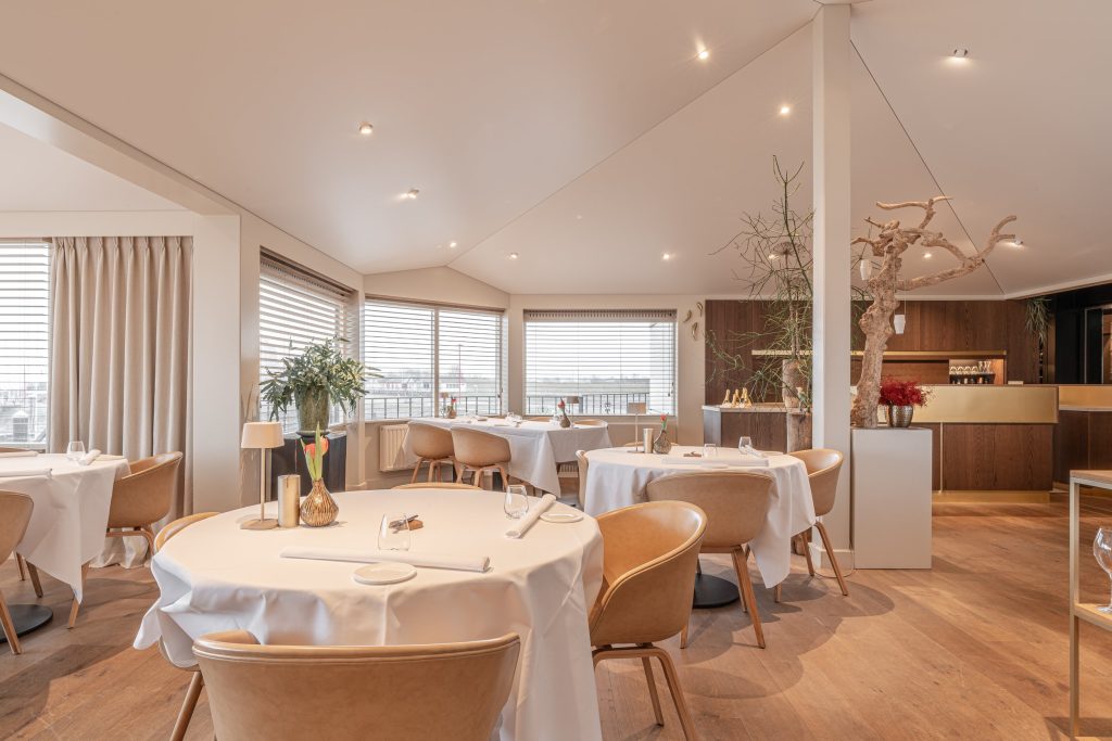 Lichte eetzaal met ronde, wit gedekte tafels en uitzicht door grote ramen.