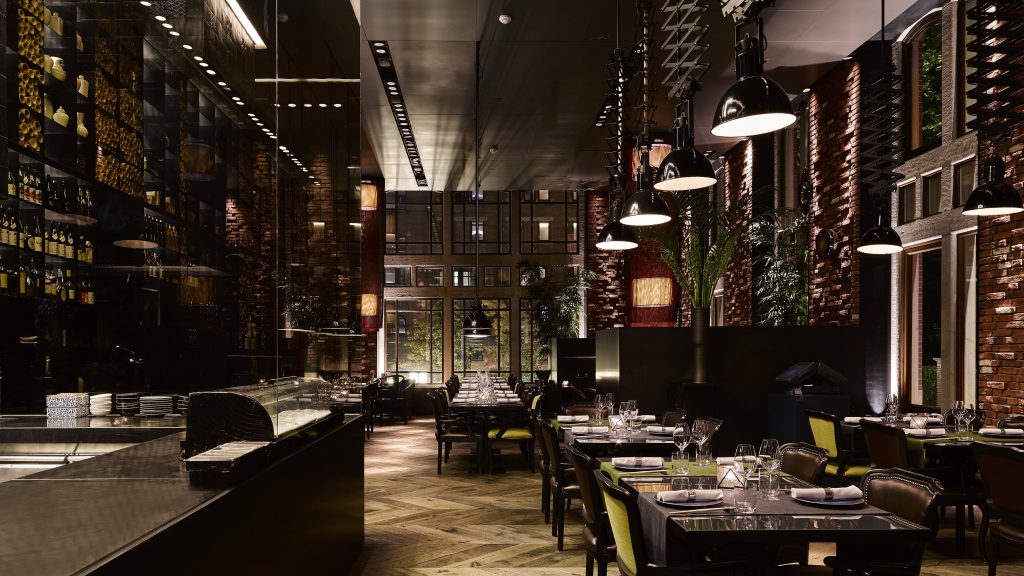 Donkere, hoge restaurantzaal met grote ramen en gedekte tafels.