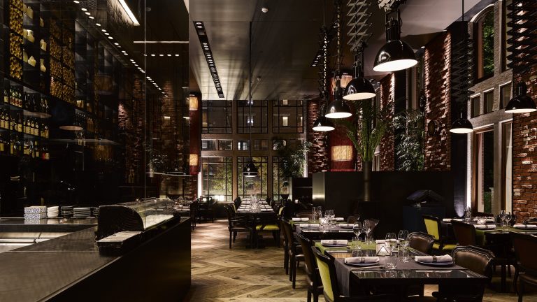 Donkere, hoge restaurantzaal met grote ramen en gedekte tafels.