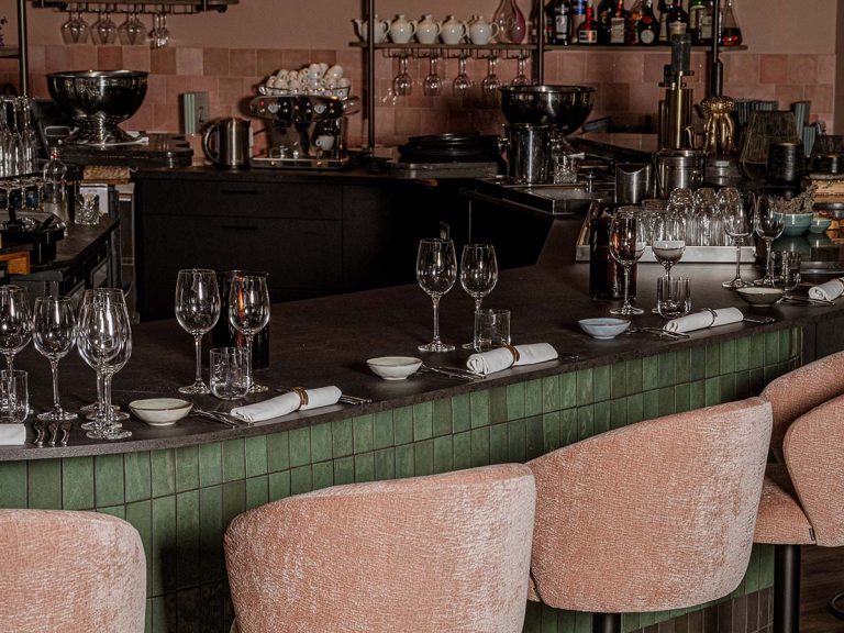 Restaurantbar met groene tegels, roze barkrukken en wijnglazen.