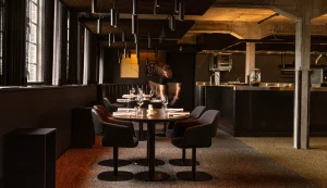 Restaurantinterieur met gedekte ronde tafels, open keuken en houten balken.