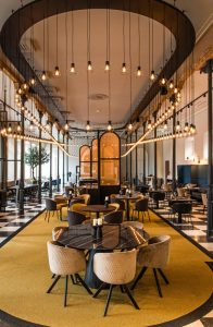 Langgerekte restaurantzaal met hoge ramen en ronde tafels.