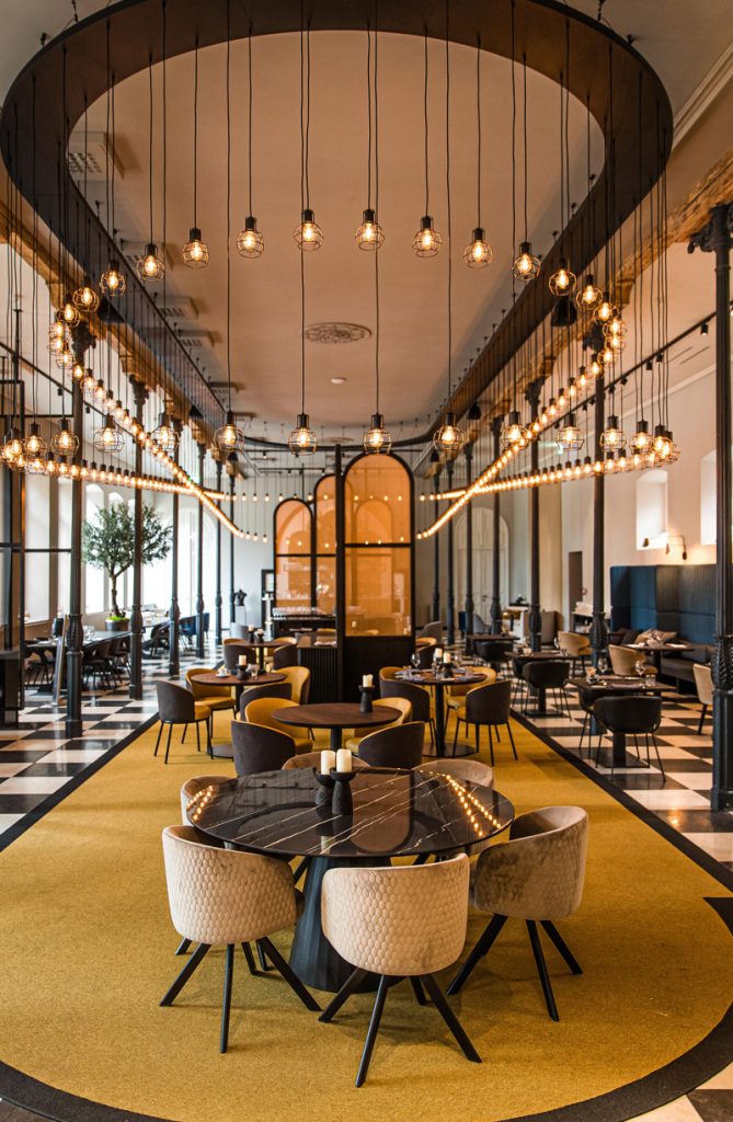 Langgerekte restaurantzaal met hoge ramen en ronde tafels.