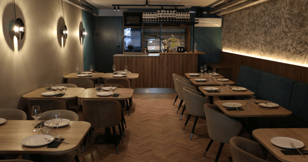 Restaurantinterieur met ronde tafels, lange wandbank en bar achterin.