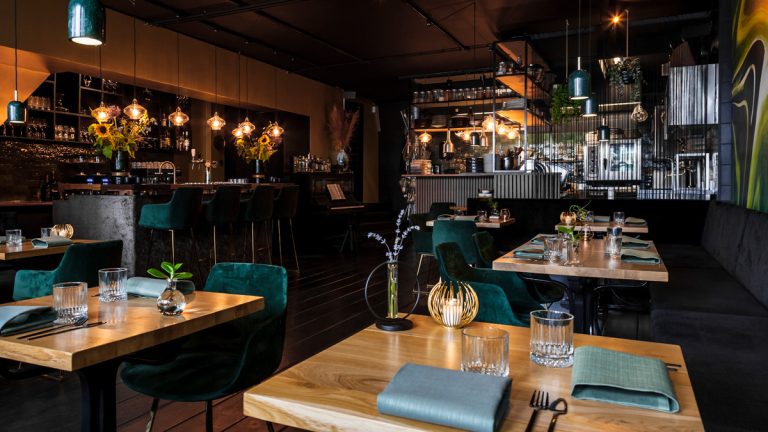 Restaurantzaal met groene stoelen, houten tafels en bar.