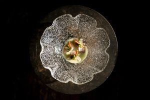 Klein culinair gerecht op een glazen, bloemvormig bord tegen zwarte achtergrond.