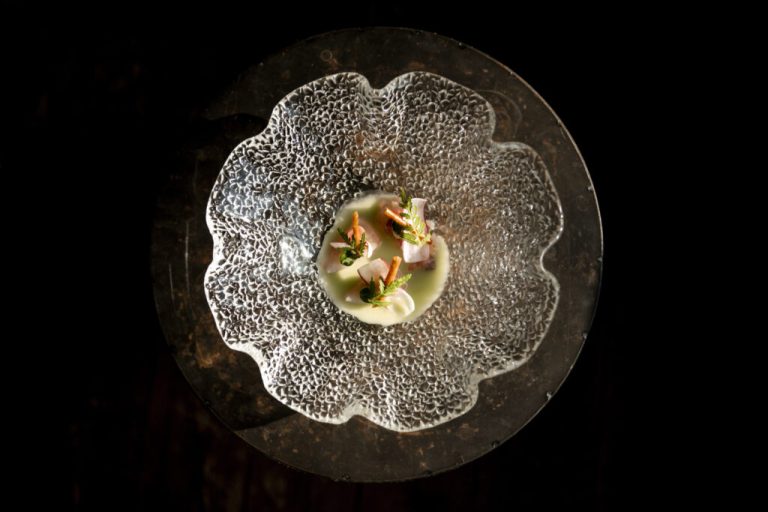 Klein culinair gerecht op een glazen, bloemvormig bord tegen zwarte achtergrond.