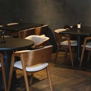 Rustig donker restaurantinterieur met houten stoelen, ronde tafels en visgraatvloer.