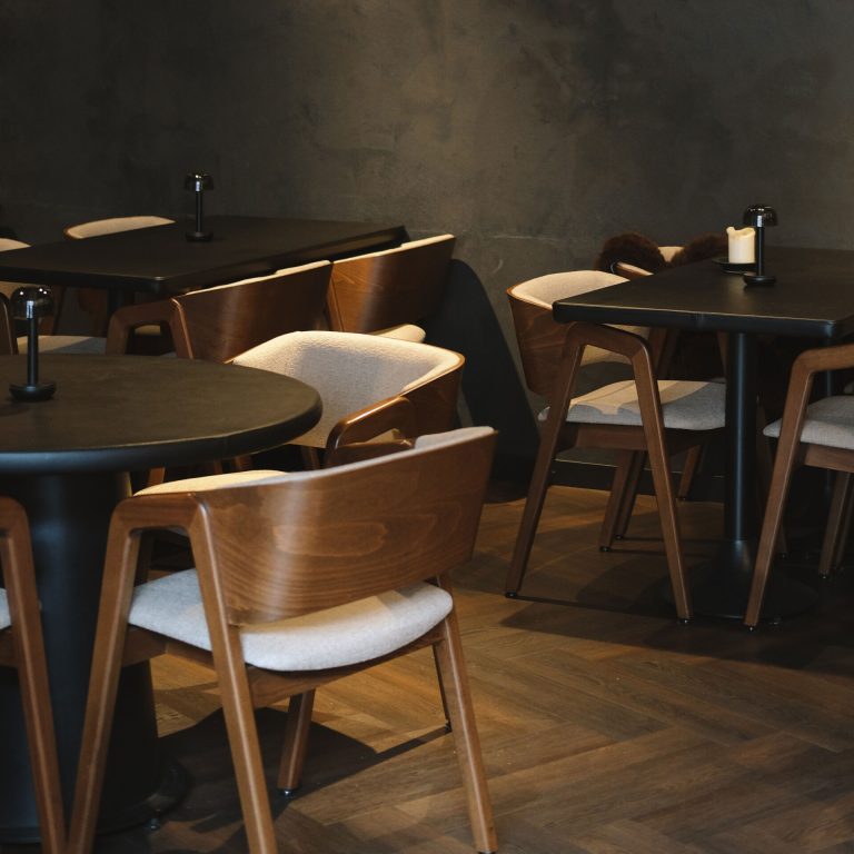 Rustig donker restaurantinterieur met houten stoelen, ronde tafels en visgraatvloer.