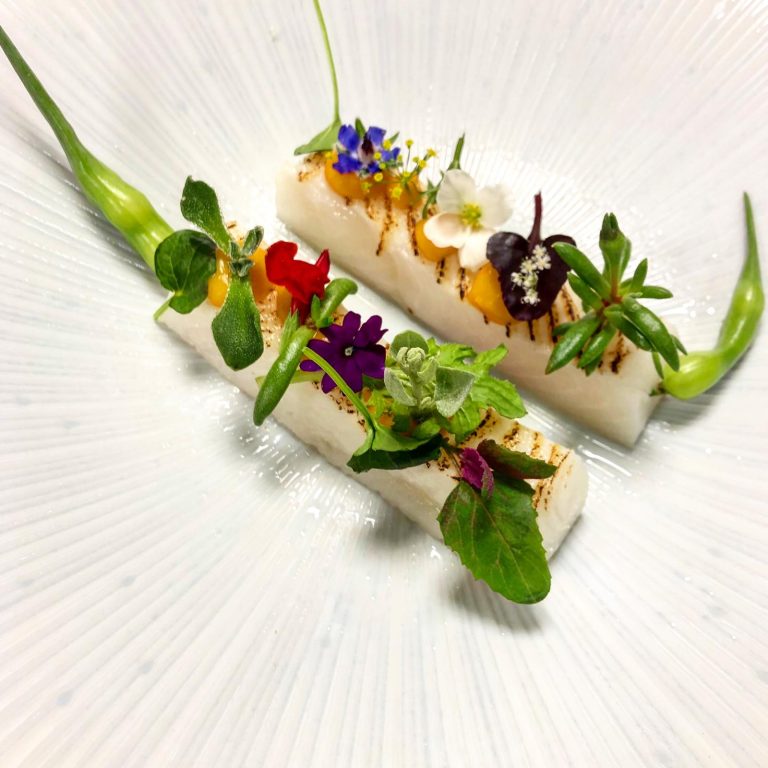 Twee opgemaakte asperges met groene en bloemige garnituur op wit bord.