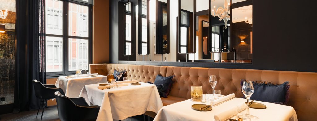 Restaurantzaal met gecapitonneerde bank, witte tafellinnen en donkere wandbetimmering.