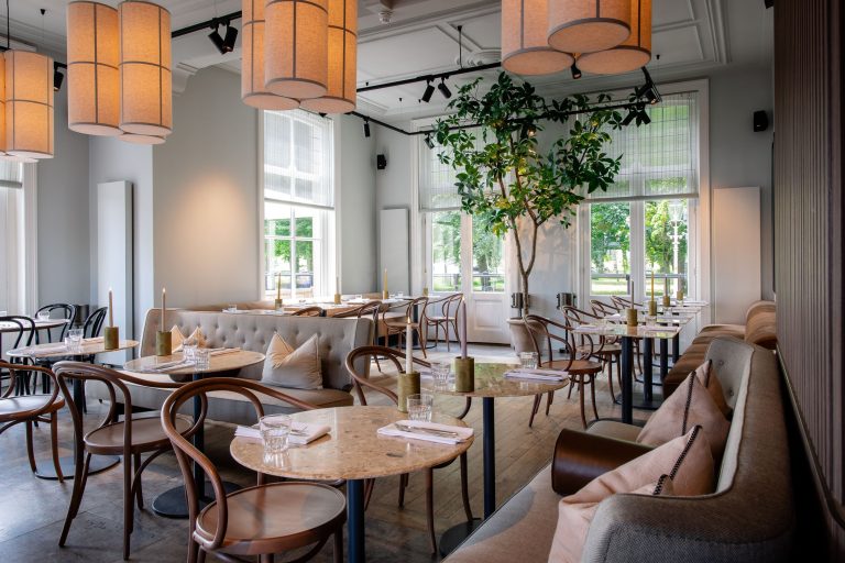 Lichte restaurantzaal met ronde tafels, beklede banken, hoge ramen en een boom.