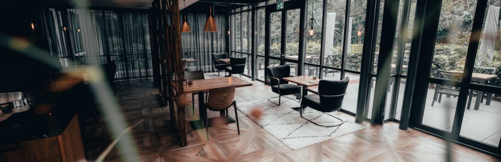 Brede eetzaal met tafels en grote raampartijen.