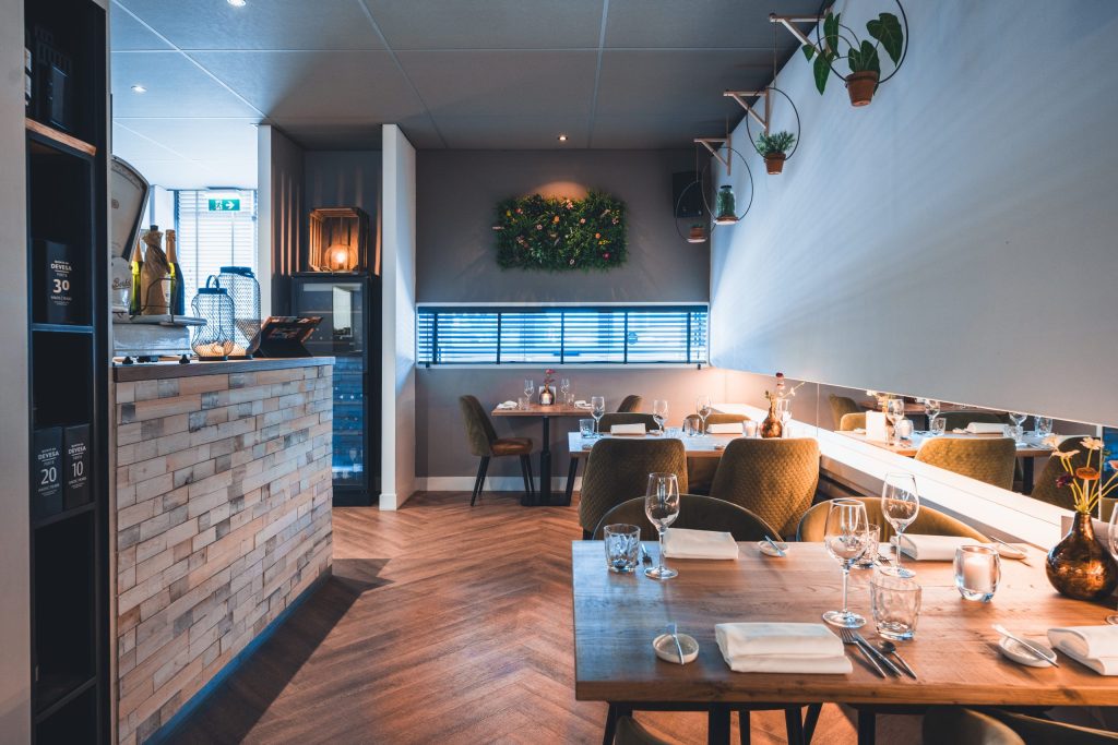 Restaurantzaal met open keuken en bar, houten vloer en gedekte tafels.
