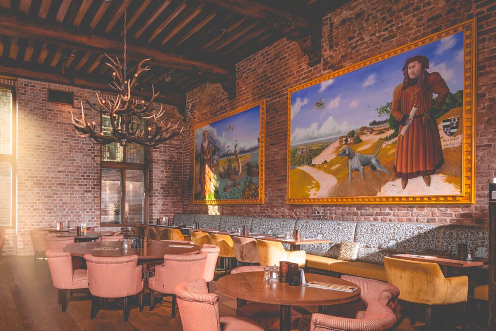Restaurantzaal met bakstenen muren, schilderijen en warmgekleurde stoelen.