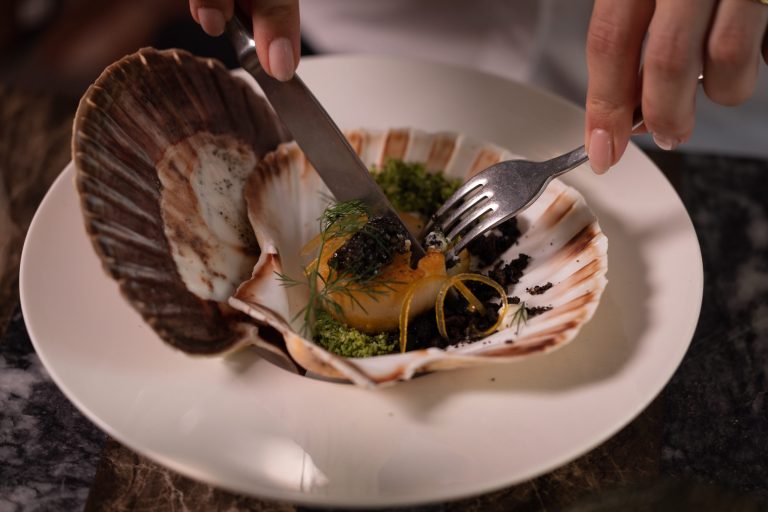 Pasta met een grote schelp op een wit bord; handen met bestek zichtbaar.