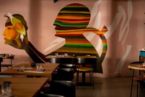 Restaurantzaal met tafels en een kleurrijke abstracte muurschildering.