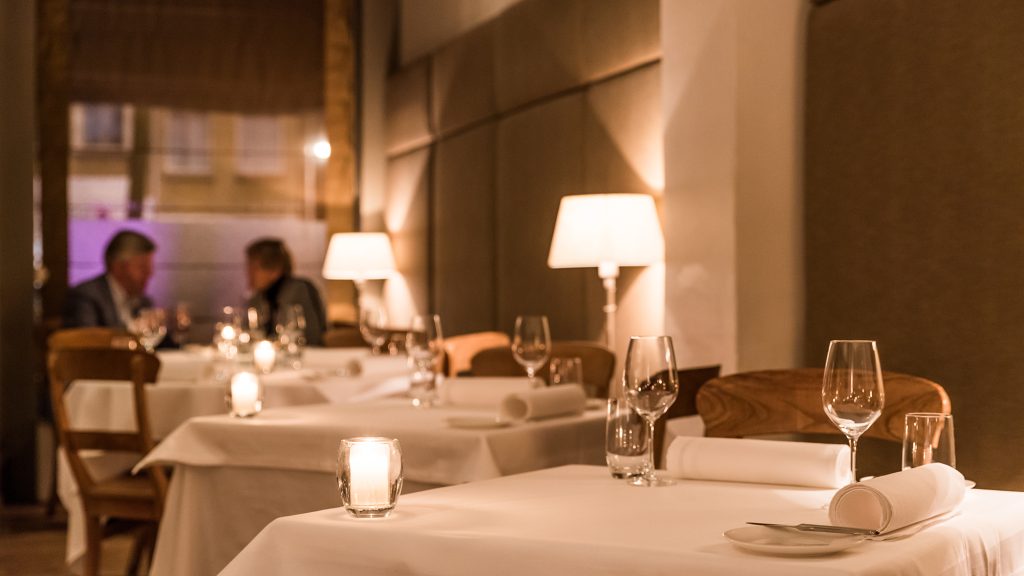 Warm restaurantinterieur met witte tafelkleden, tafellampen en gedekte tafels.