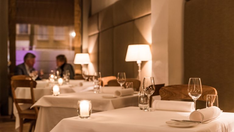 Warm restaurantinterieur met witte tafelkleden, tafellampen en gedekte tafels.