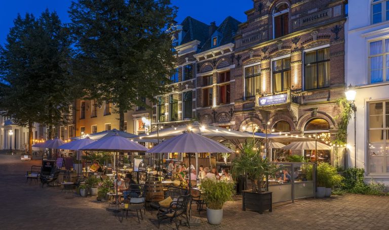Verlicht restaurantpand en terras aan het plein in de avond.