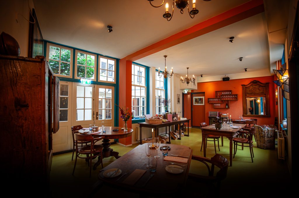 Warme eetzaal met houten tafels, grote ramen en rode wand.
