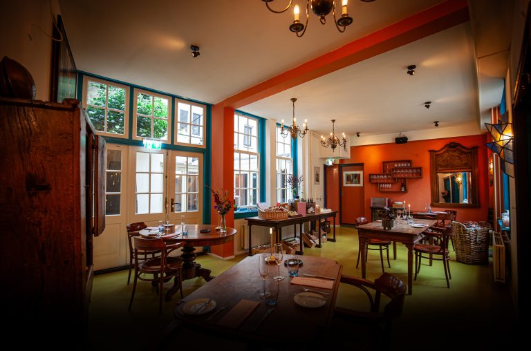 Warme eetzaal met houten tafels, grote ramen en rode wand.