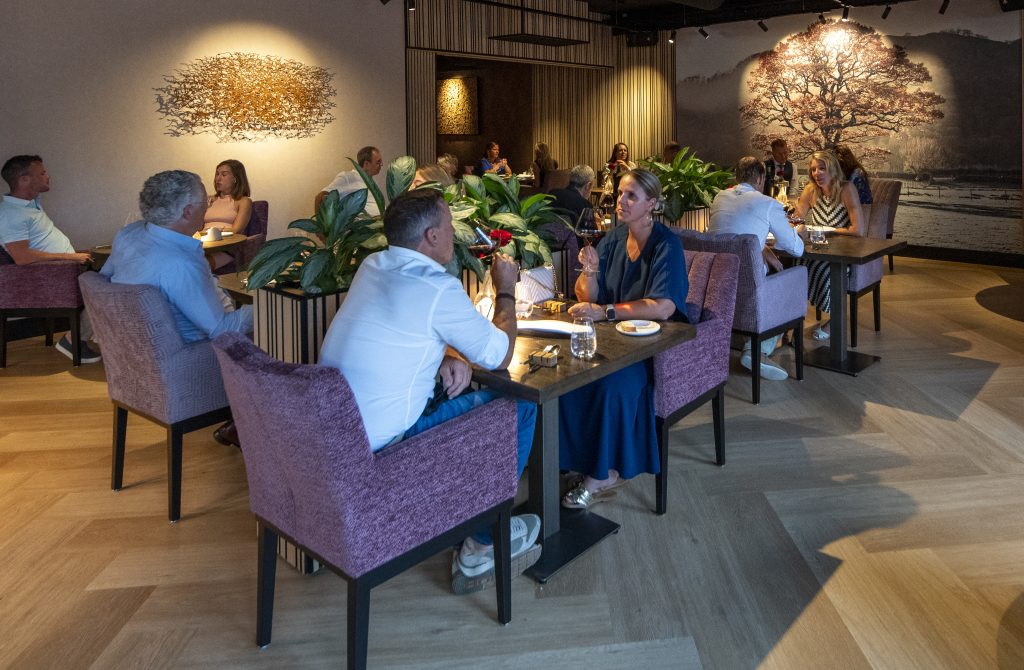 Gasten in restaurantzaal met gekleurde stoelen en ronde tafels.