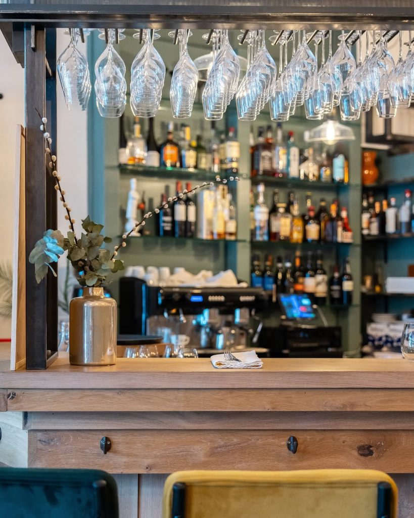 Houten bar met hangende wijnglazen, flessen, koffieapparaat en barkrukken.